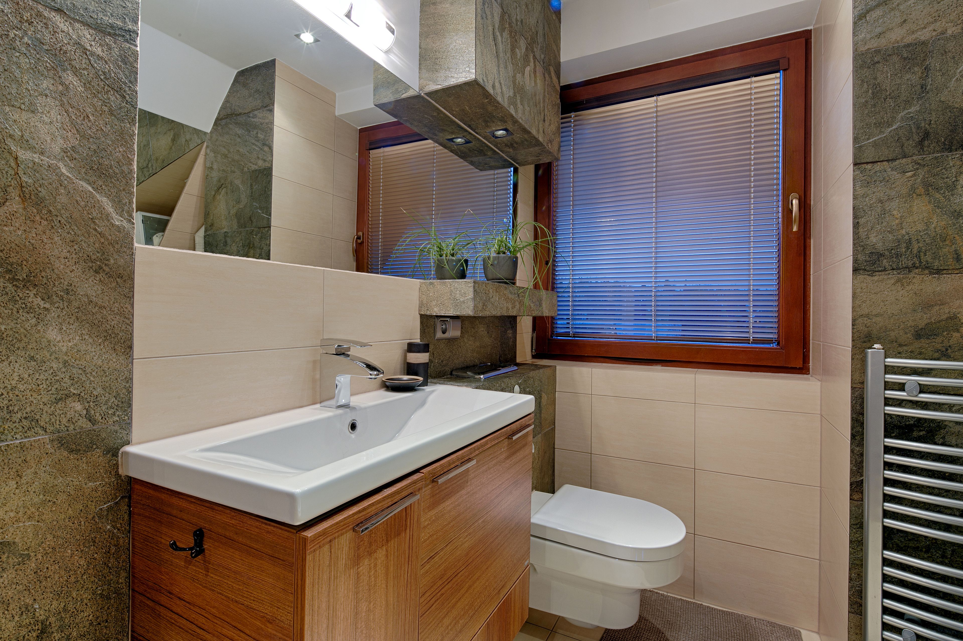 Un baño moderno con lavamanos de encimera de granito, muebles de madera, inodoro y ventana con persianas.
