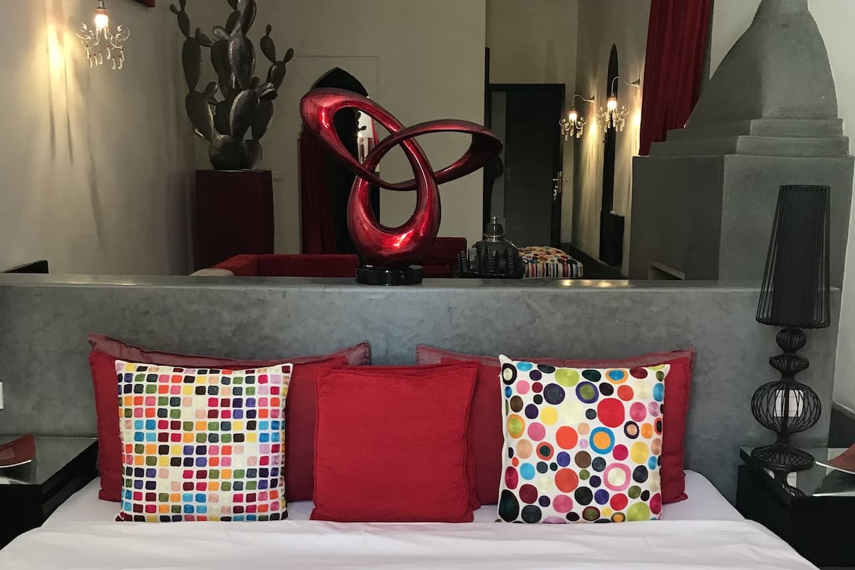 A hotel room with a bed, red pillows, a red abstract sculpture, a mirror, and a red curtain.