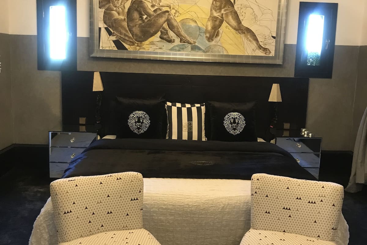A hotel room with a large bed, two bedside tables, a headboard with decorative elements, and two armchairs.