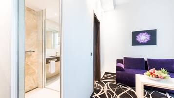 A modern hotel room with a glass shower, a purple sofa, and a fruit bowl on a white table.