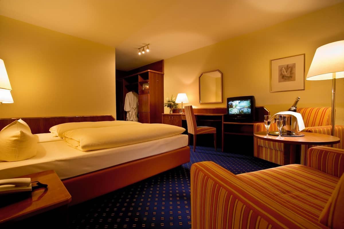 A hotel room with a bed, a desk with a television, a chair, and a small table with a bottle and glasses.
