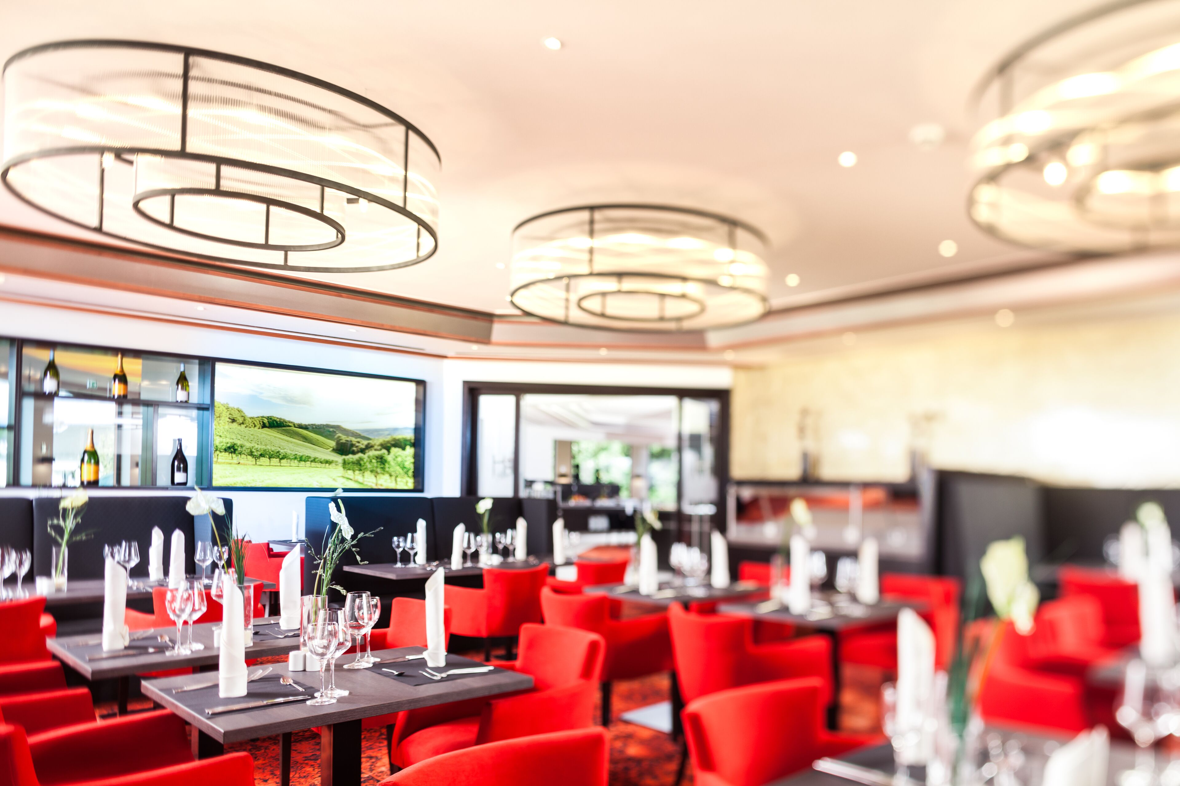 Um restaurante com cadeiras e mesas vermelhas, luminárias modernas e vista para um vinhedo por grandes janelas.