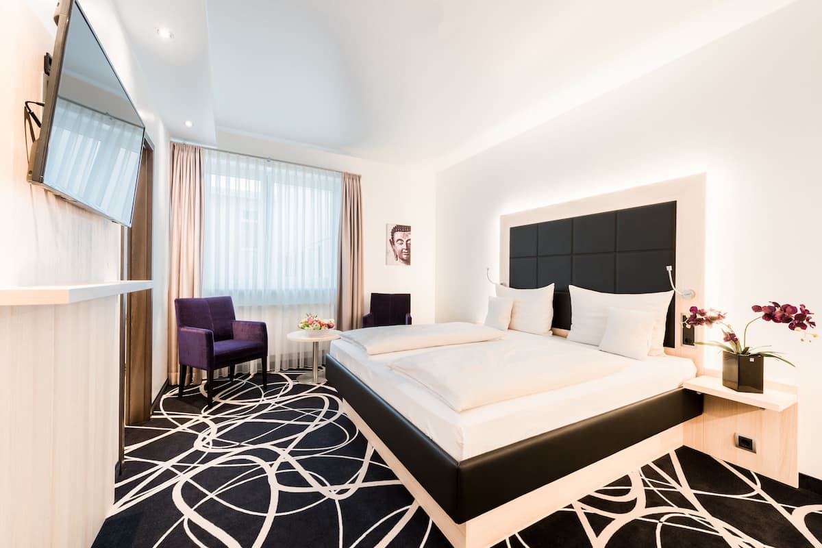 A modern hotel room with a large bed, a flat-screen TV mounted on the wall, a small round table with a vase of flowers, and a chair.