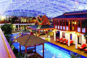 An indoor resort with a swimming pool, a thatched-roof structure, and a traditional building with red chairs. - Sieben Welten Hotel & Spa Resort (Kuenzell)