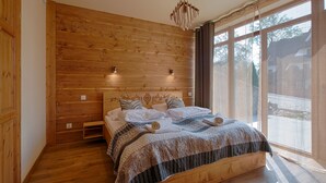 A bedroom with a wooden headboard, a bed with a patterned bedspread, two bedside lamps, and a window with a view of the outside. - Tatrzańskie Echo Domki Regionalne (Zakopane)