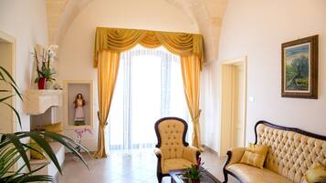 A spacious living room with a high arched ceiling, a large window with yellow curtains, a plush sofa, a chair, and a small table with a plant.