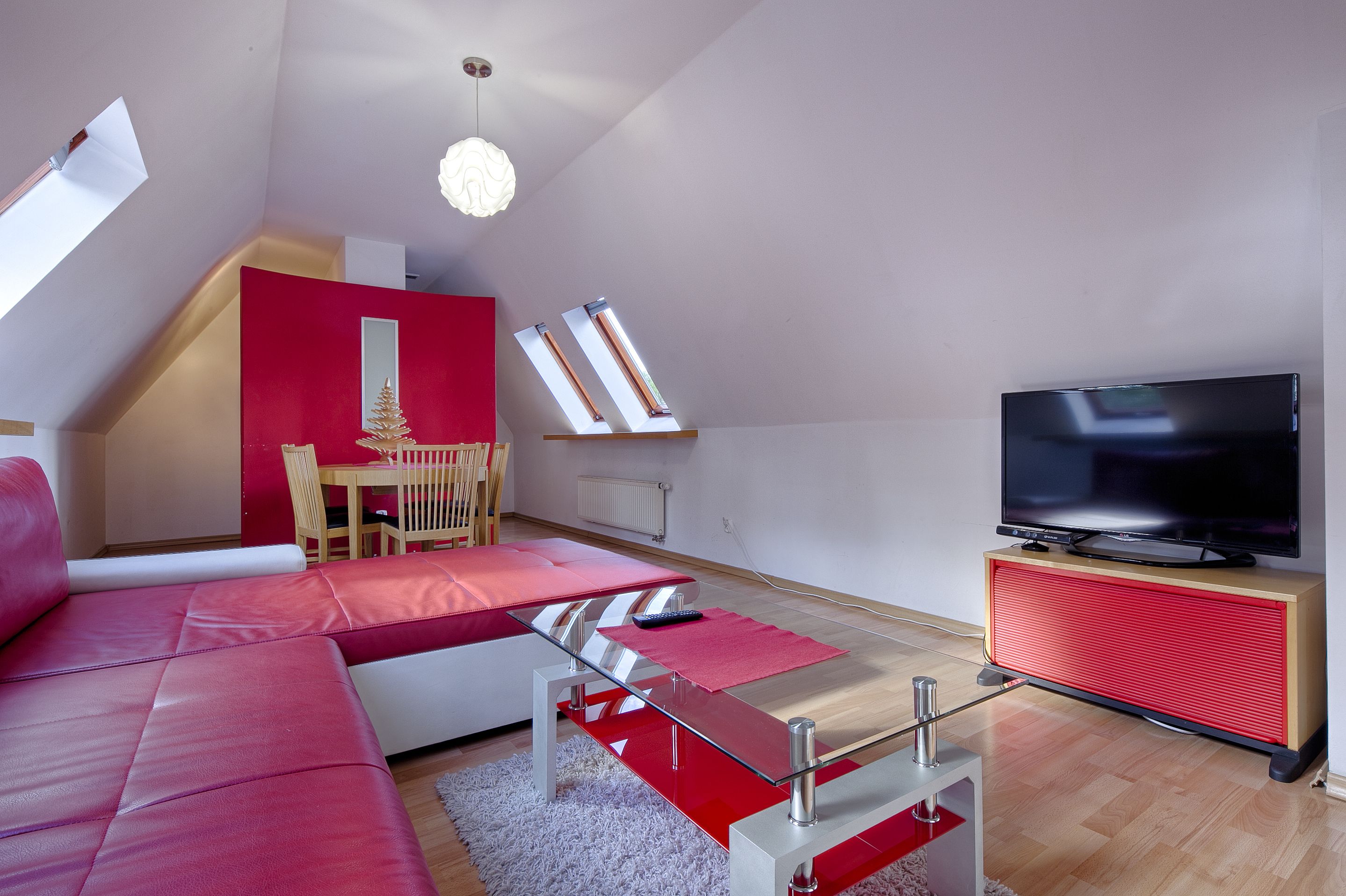 A modern living room with a red sofa, a glass coffee table, a TV on a red stand, and a dining area with a table and chairs.