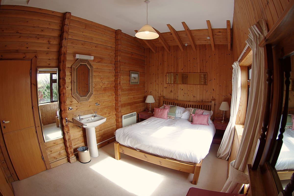A wooden cabin bedroom with a bed, bedside table, lamp, and a window with curtains.