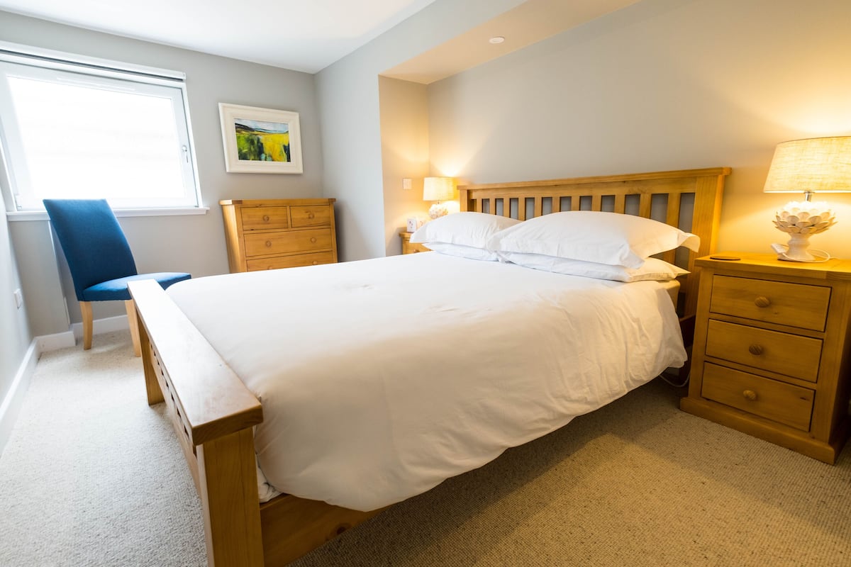 A bedroom with a bed, a wooden nightstand, a lamp, a chair, a dresser, and a window.