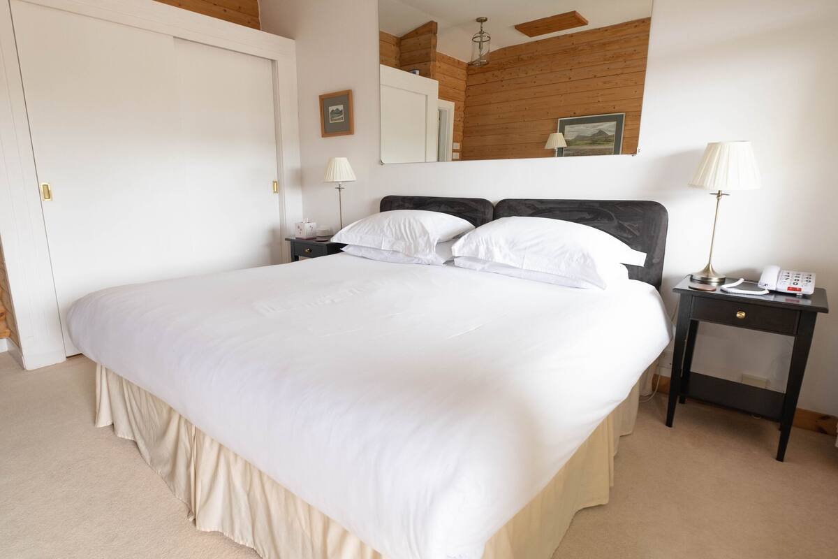 A neatly made bed with white linens in a room with wooden walls and a wooden ceiling. There is a bedside table with a lamp and a phone.