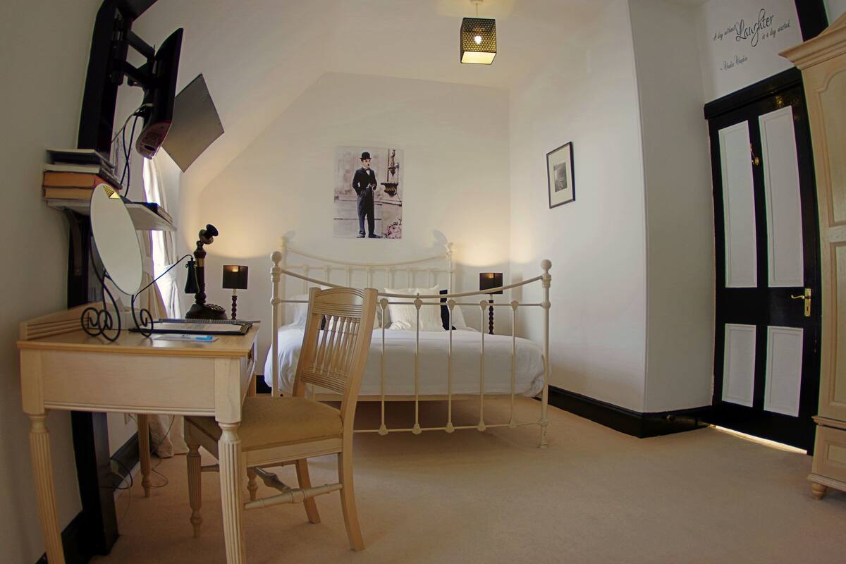 A bedroom with a bed, a desk with a chair, a desk lamp, a bookshelf, a wall-mounted TV, and a door with a black and white design.