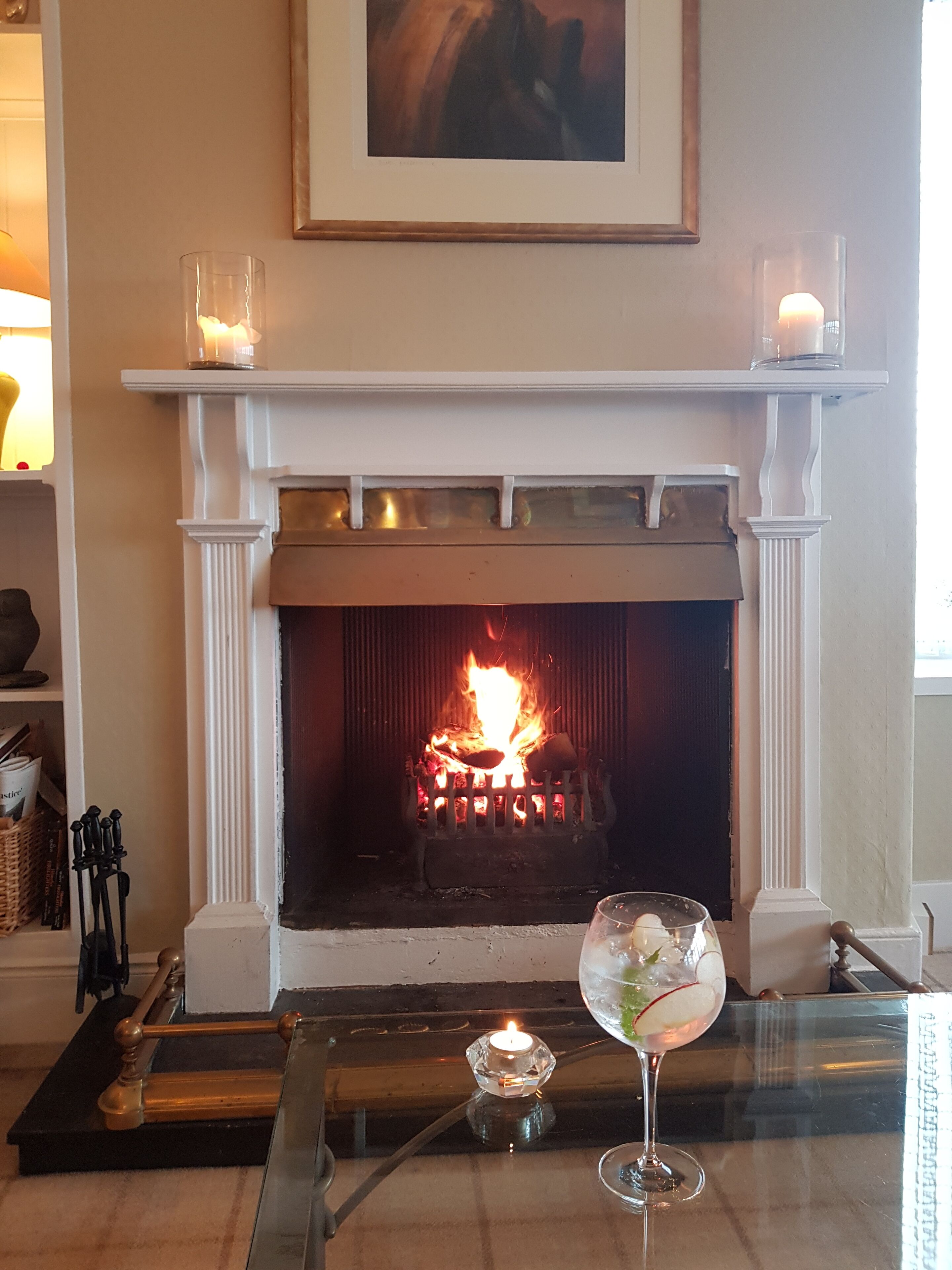 Una chimenea encendida, un cuadro colgado encima y una mesa de vidrio con una vela y un vaso de agua.