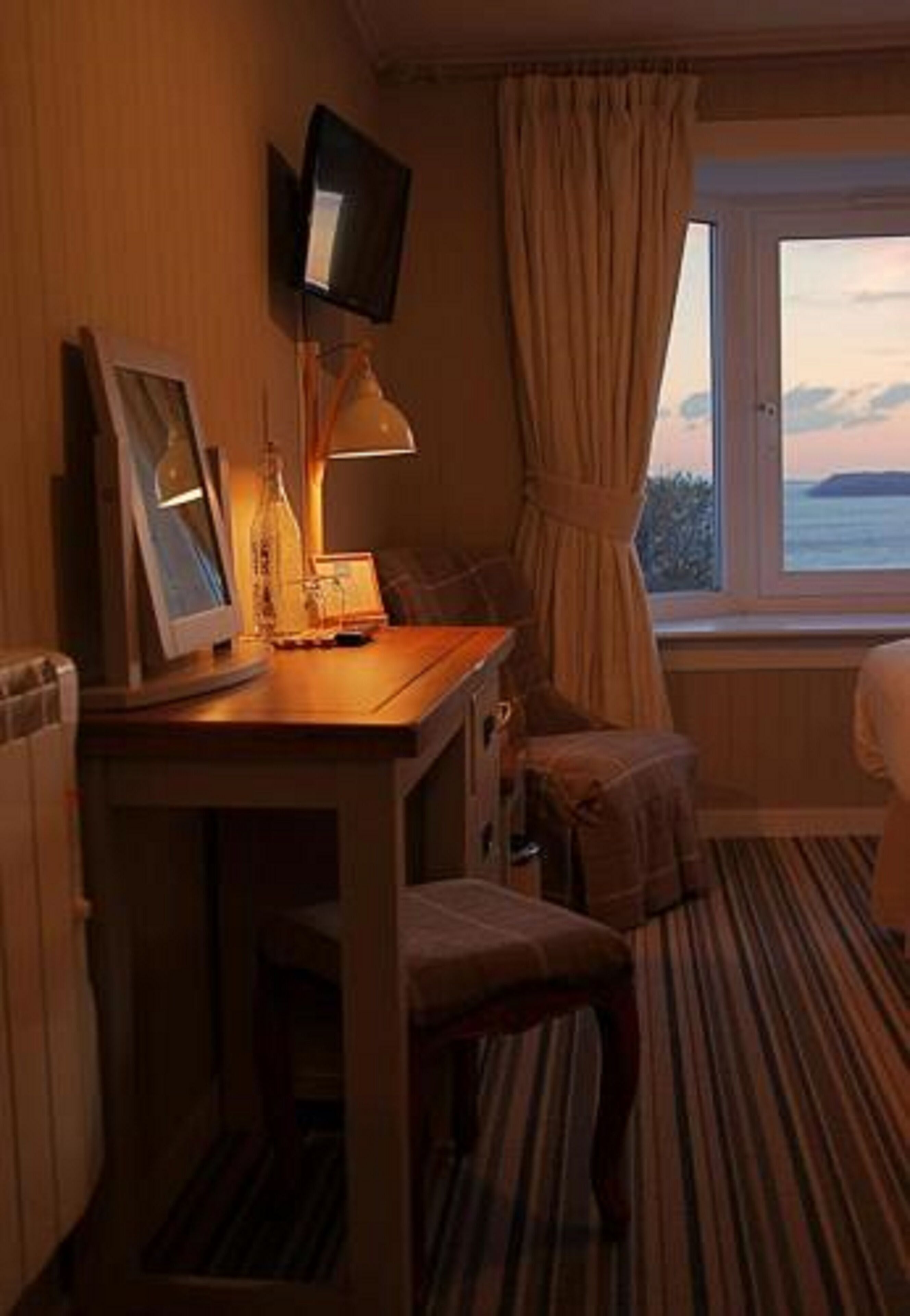 A hotel room with a wooden desk, a television mounted on the wall, a window with curtains, and a view of the sea.