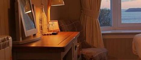 A hotel room with a wooden desk, a television mounted on the wall, a window with curtains, and a view of the sea.
