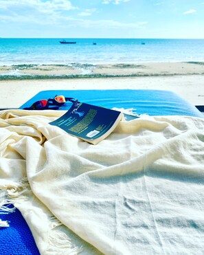 A beach scene with a book and sunglasses on a blue mat, a white cloth, and the ocean in the background.
