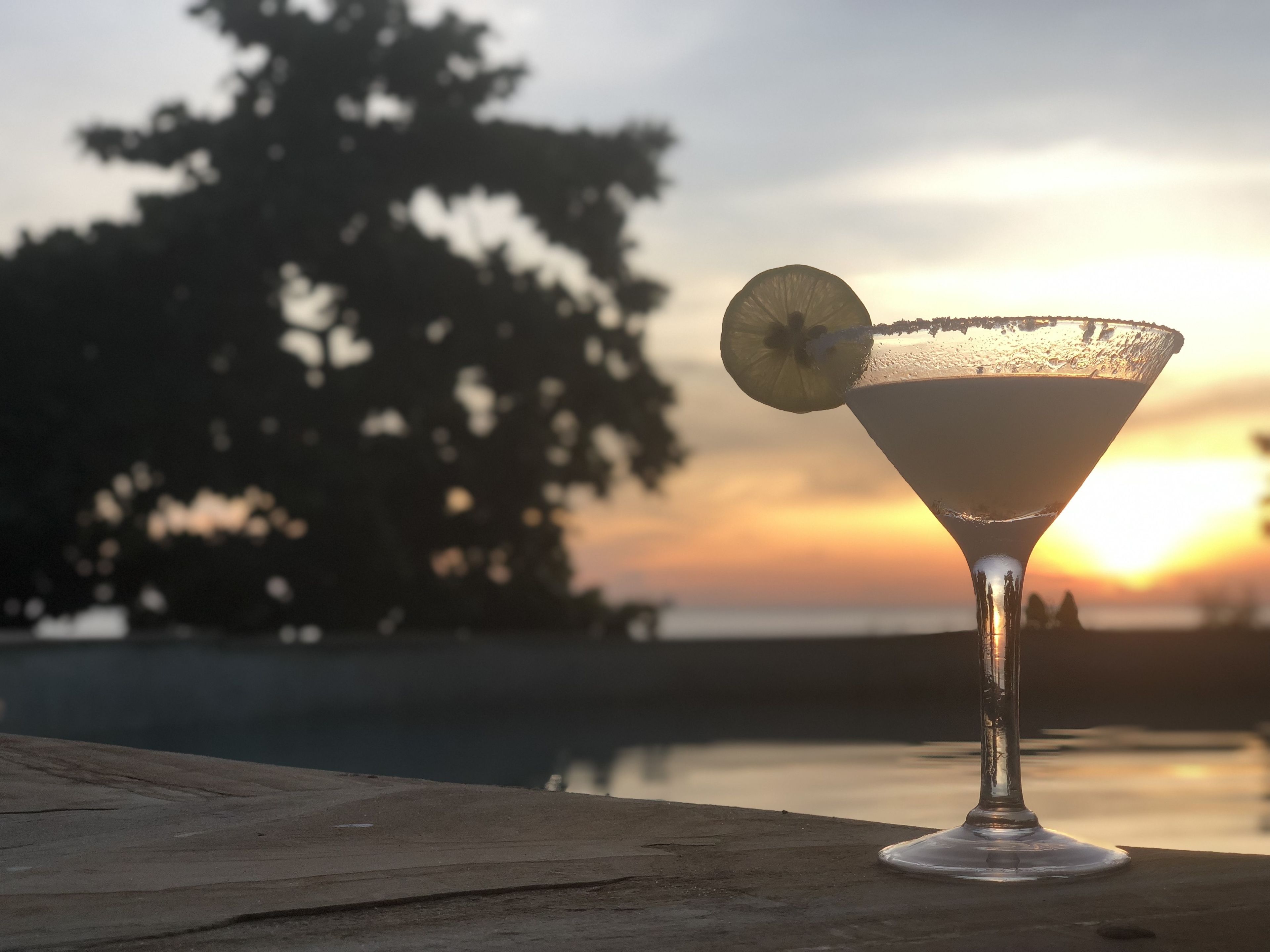 A martini glass with a lemon slice on a wooden surface, set against a sunset background.