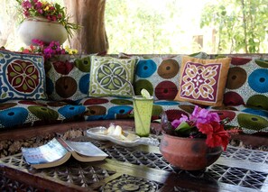 A cozy seating area with colorful cushions, a table with a drink, a book, and a flower pot.