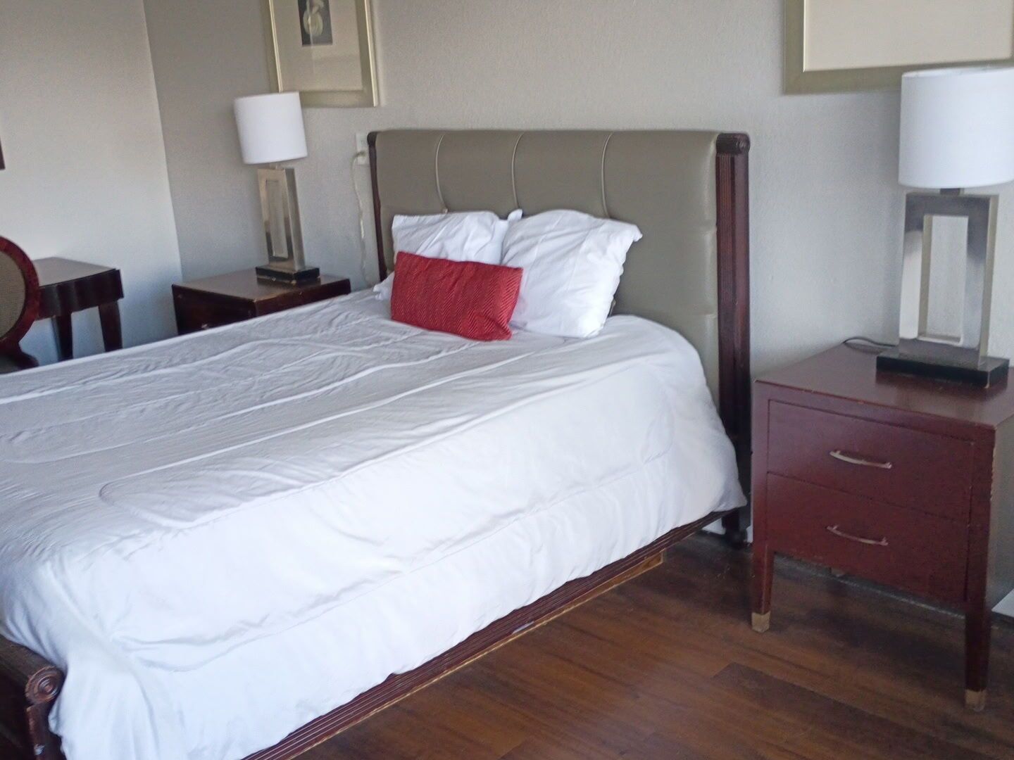 Een keurig opgemaakt bed met wit beddengoed en een rode kussen, een houten nachtkastje met lamp en een houten dressoir met laden.