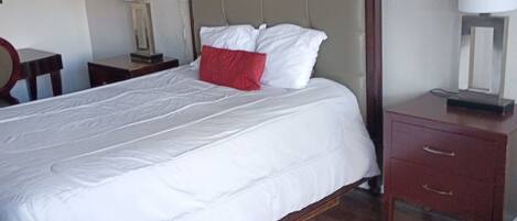 Een keurig opgemaakt bed met wit beddengoed en een rode kussen, een houten nachtkastje met lamp en een houten dressoir met laden.