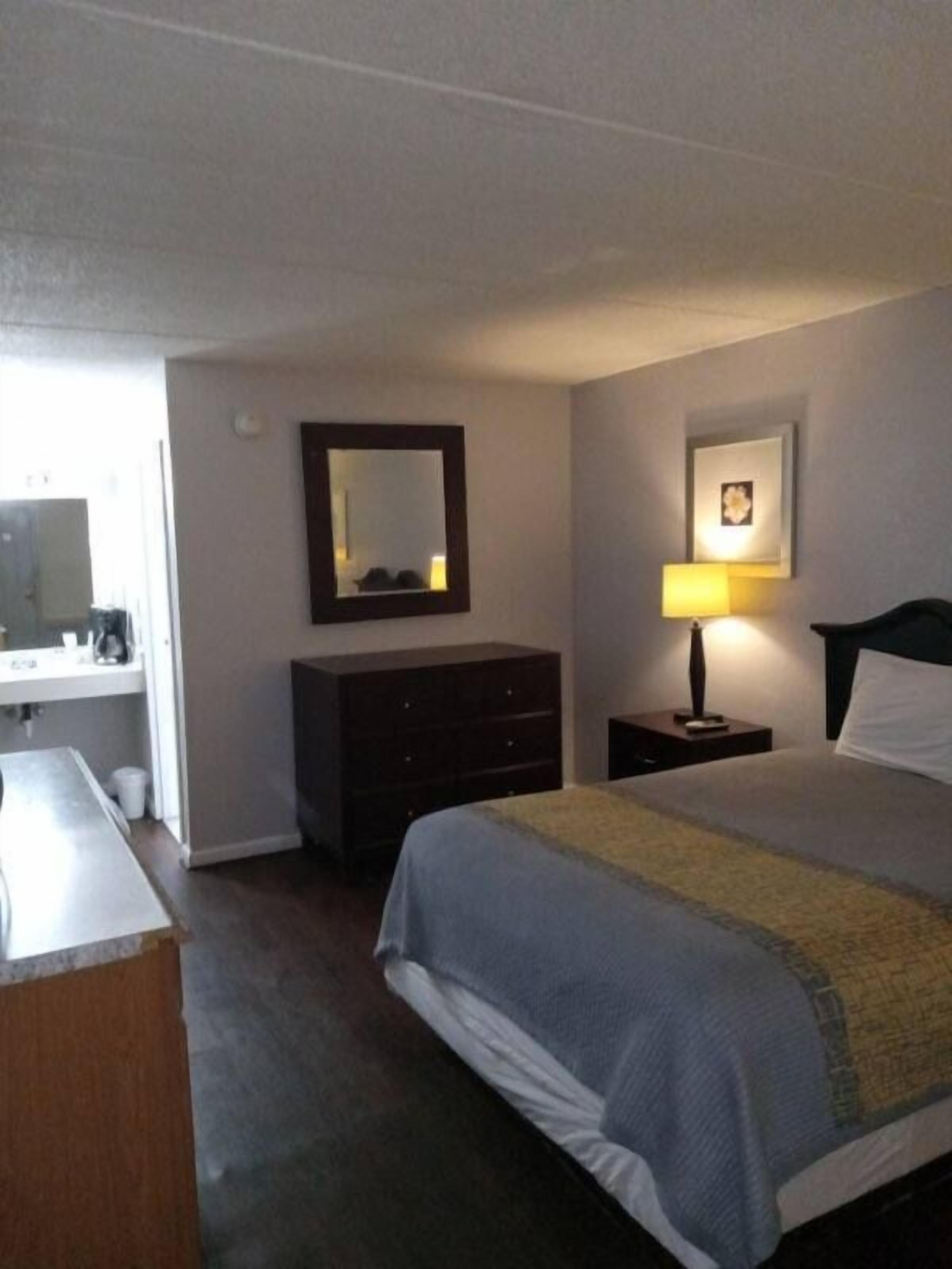 Une chambre d’hôtel avec un lit, une commode, un miroir, une table de chevet avec une lampe et un tableau au mur.