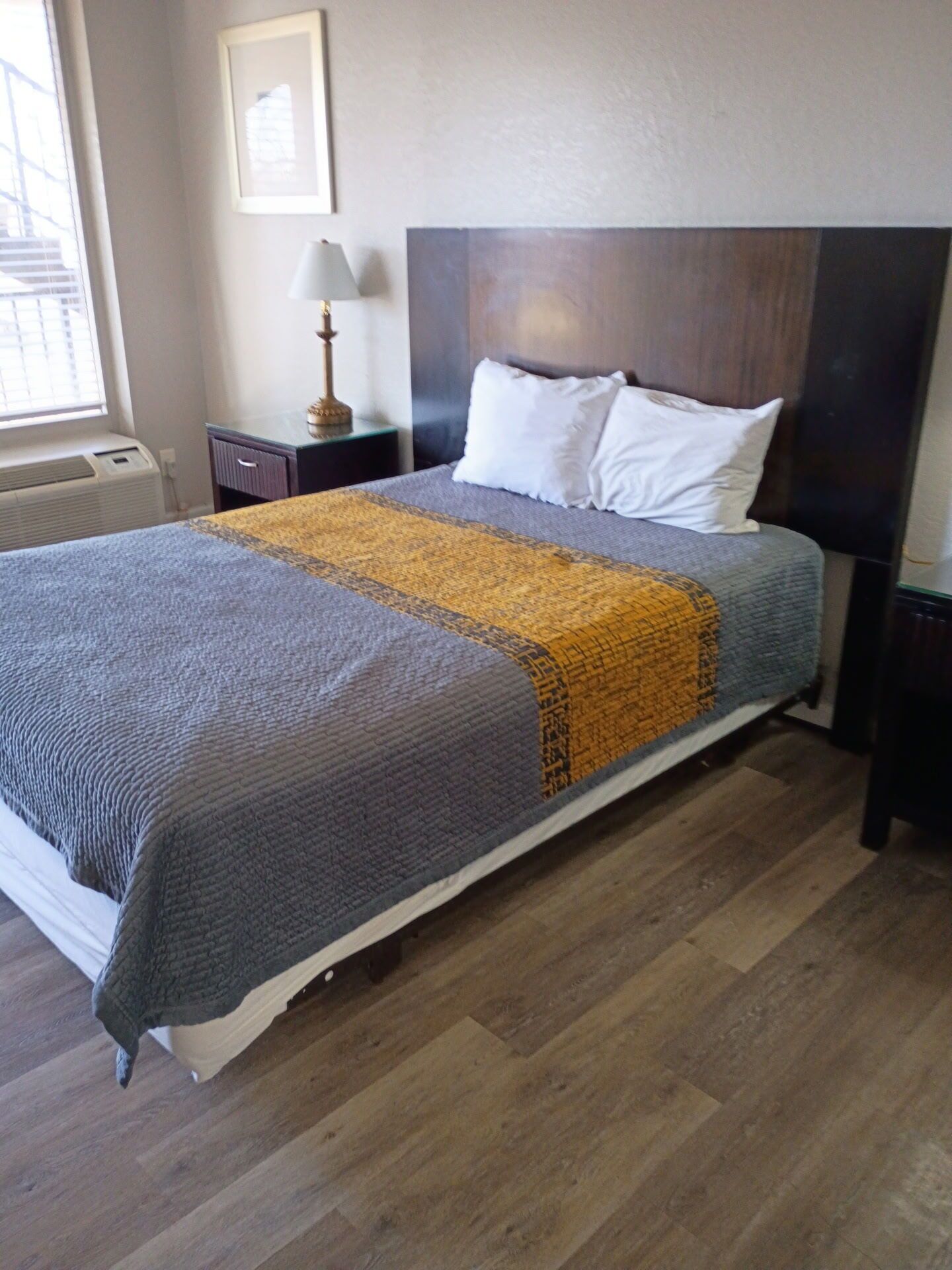 A neatly made bed with a grey and yellow blanket, a wooden headboard, and a bedside table with a lamp.