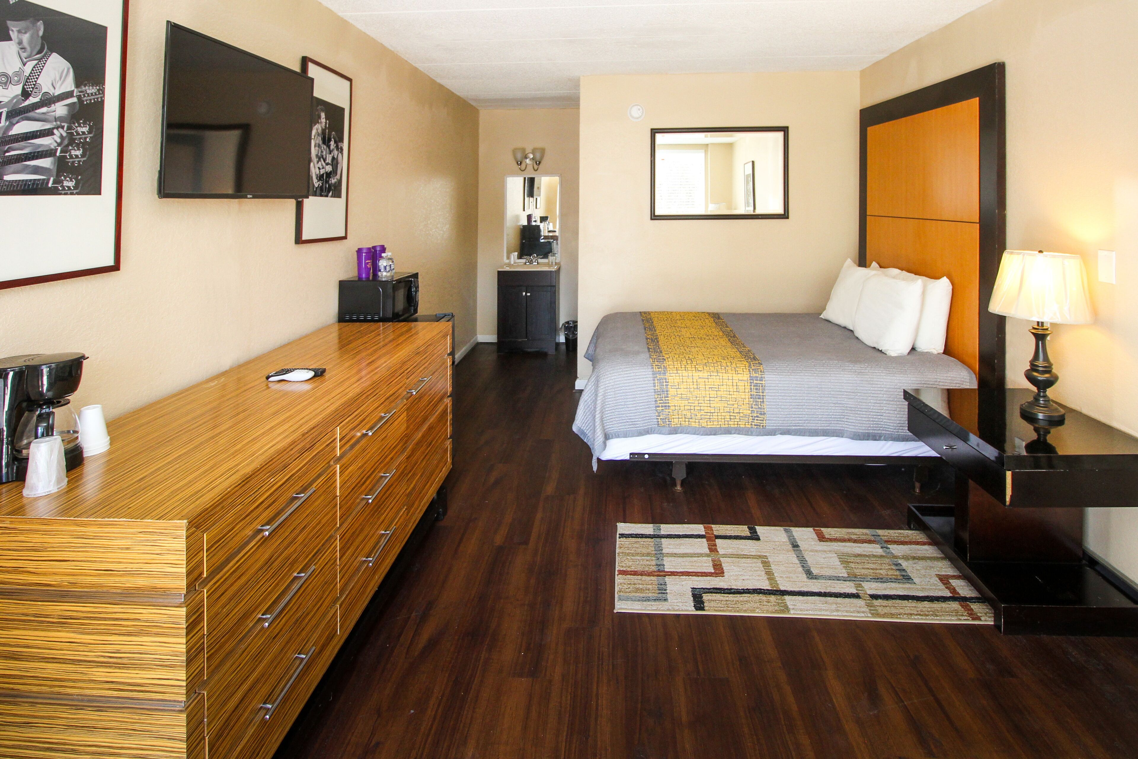 A hotel room with a wooden dresser, a bed with a gray and yellow striped bedspread, a small desk with a lamp, and a mirror above the dresser.