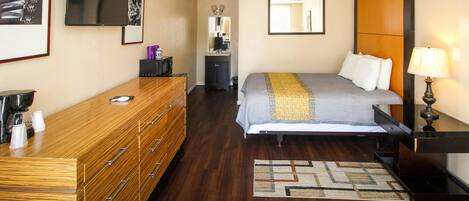 A hotel room with a wooden dresser, a bed with a gray and yellow striped bedspread, a small desk with a lamp, and a mirror above the dresser.