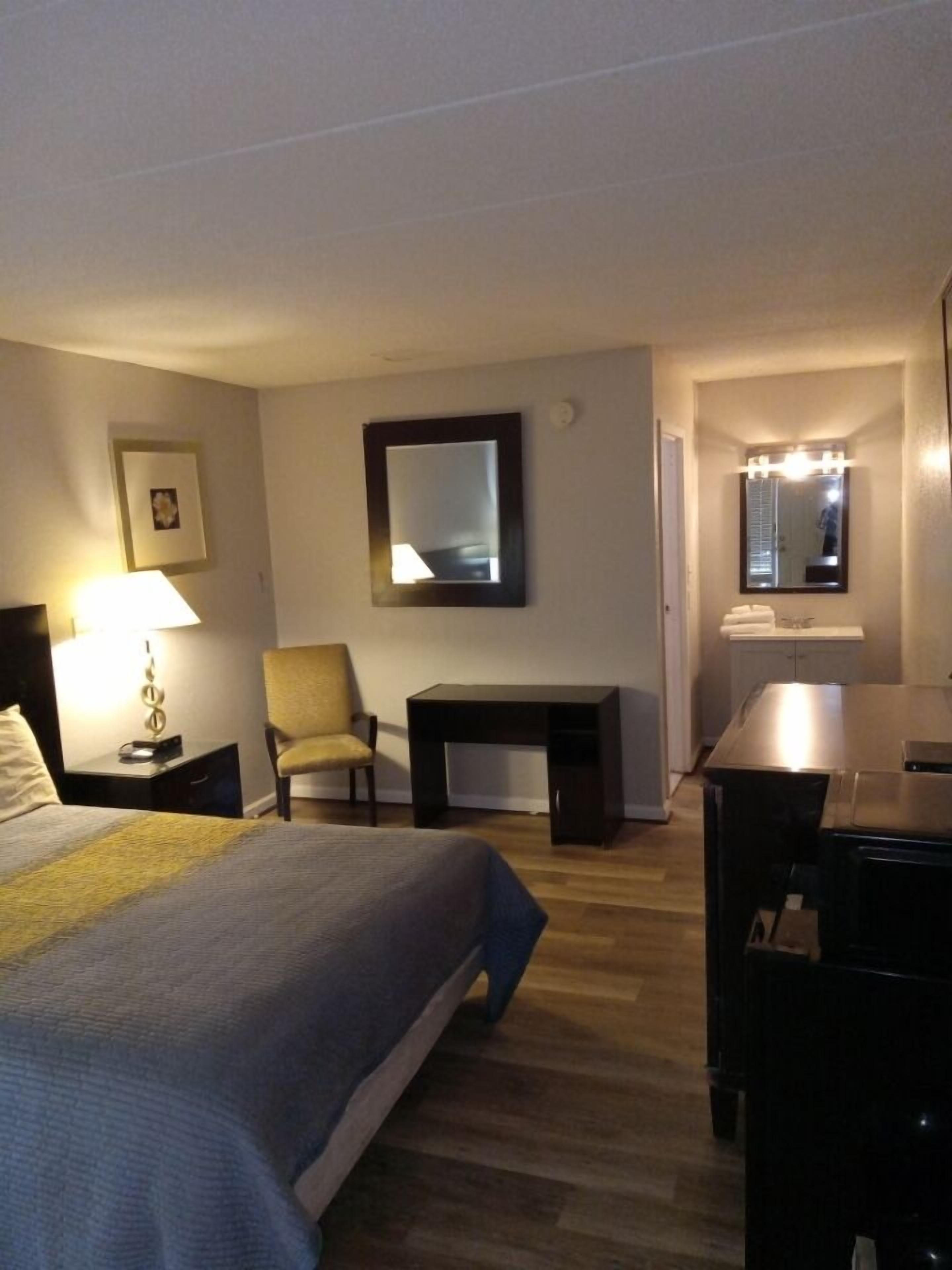 Habitación de hotel con cama, escritorio, silla, mesita, lámpara y espejo.