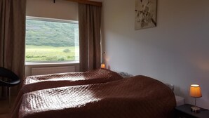 Et værelse med to senge, et vindue med gardiner, en lampe på et natbord og et billede på væggen.