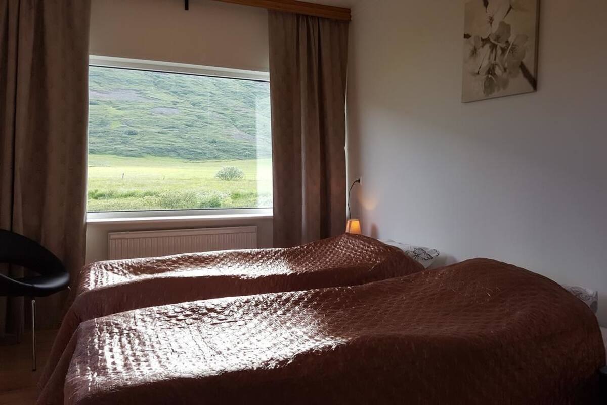 Una habitación con dos camas, una ventana con cortinas, una lámpara en una mesita de noche y un cuadro en la pared.