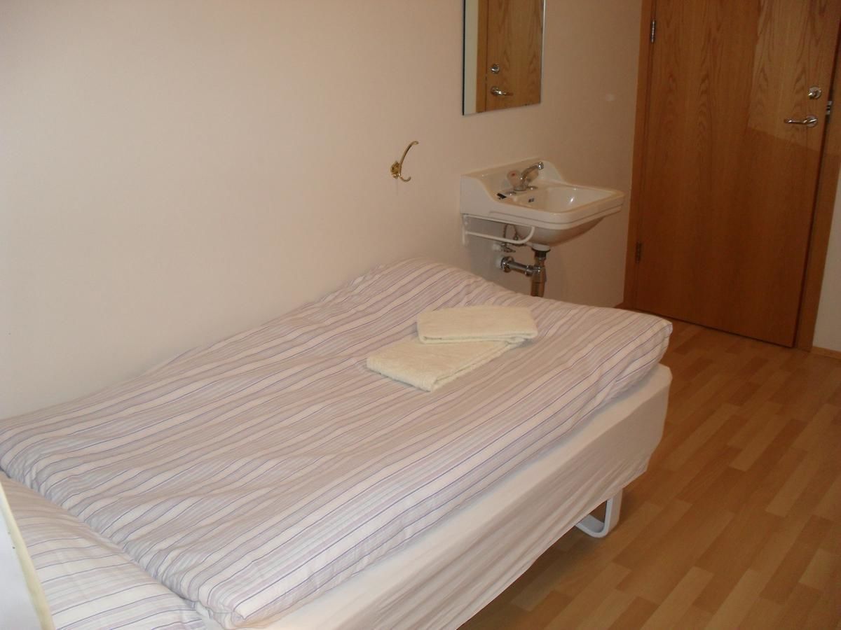 Une petite chambre avec un lit simple, un plancher en bois, un lit blanc avec une literie à rayures, un lavabo blanc et une porte en bois.