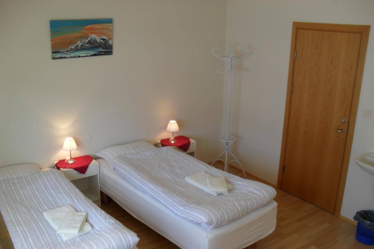 Una habitación con dos camas individuales, cada una con una mesita de noche roja y una lámpara. Hay una puerta de madera y una pared blanca con un cuadro.