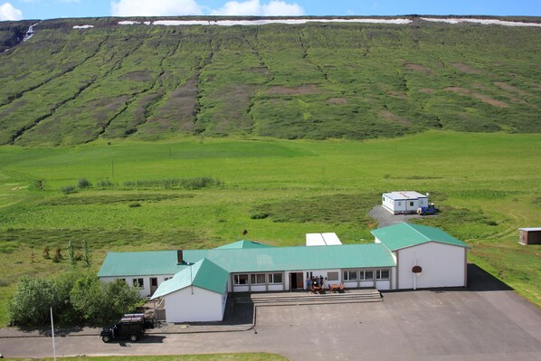 A building with a green roof and white walls, situated in a green field with a hill in the background.
