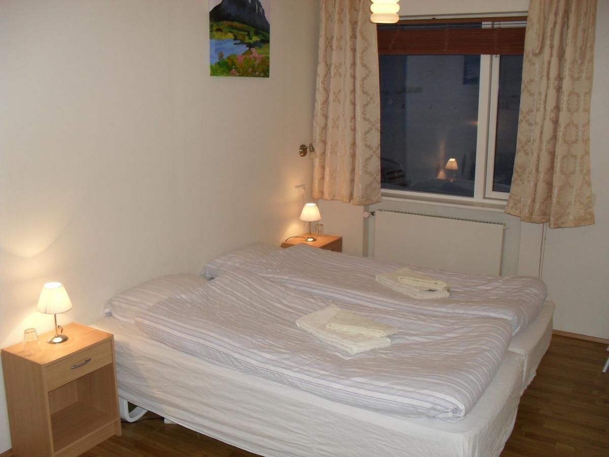 A bedroom with a double bed, two bedside tables with lamps, a window with curtains, and a painting on the wall.
