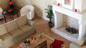 A living room with a beige sofa, a wooden coffee table with a bottle and glasses, a fireplace with a plant and decorative items, and a white chair.