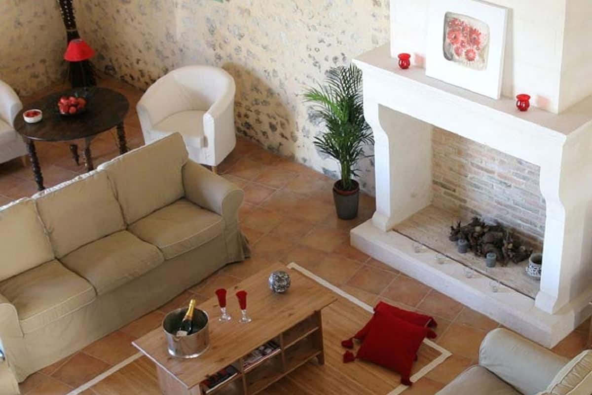 Ein Wohnzimmer mit beiger Couch, einem Holztisch mit Flasche und Gläsern, einem Kamin mit Pflanze und Dekoration sowie einem weißen Sessel.