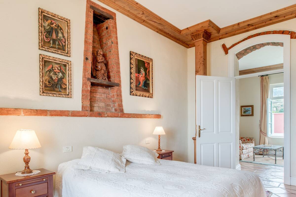 A bedroom with a bed, two bedside lamps, a brick fireplace, and a door leading to another room.