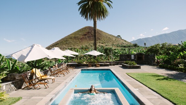 Una piscina con sillones de descanso y sombrillas, rodeada de follaje y una palmera, con una montaña al fondo.