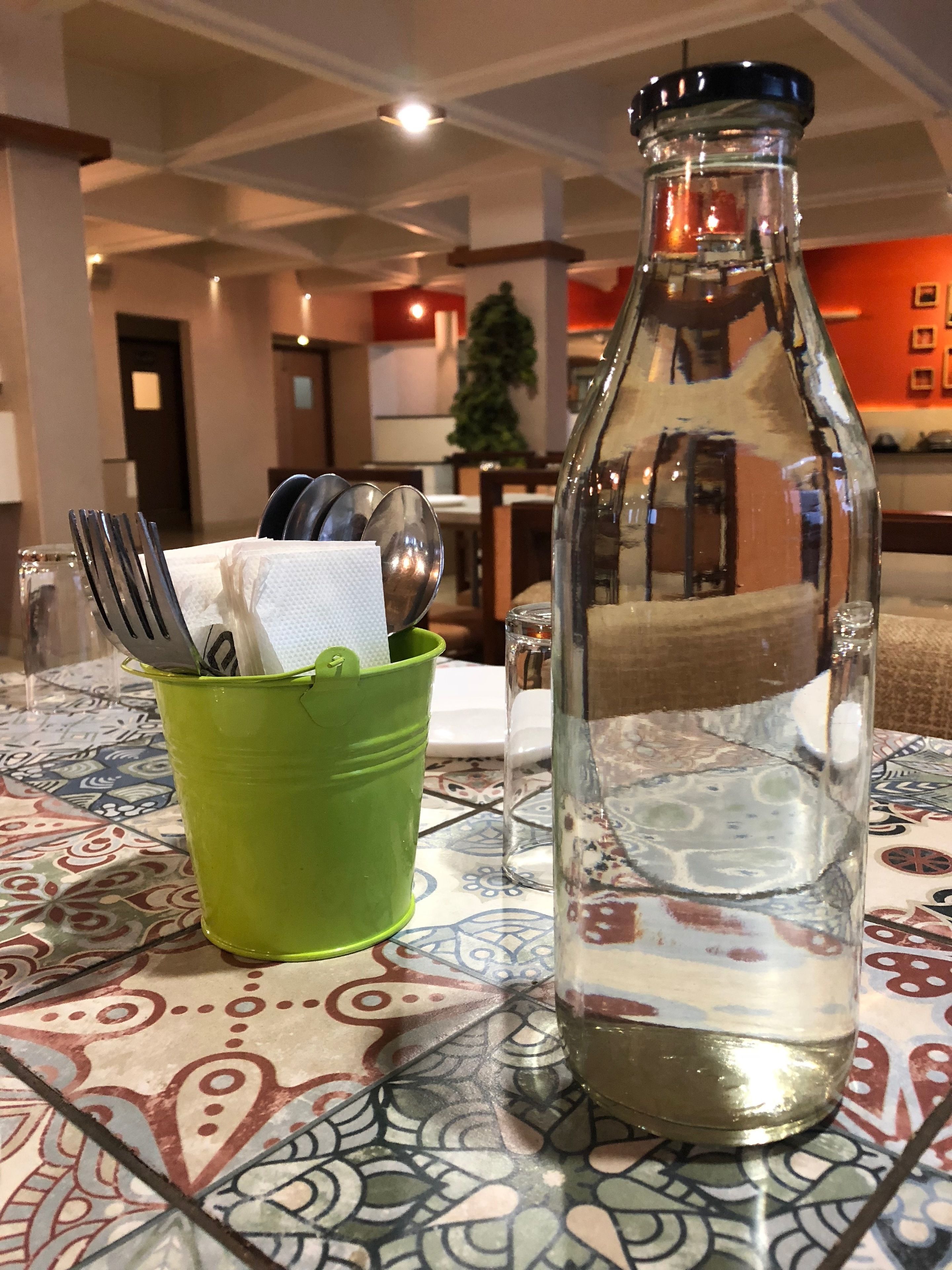A table with a patterned tablecloth, a green cup holding utensils, and a clear bottle with a metal cap.