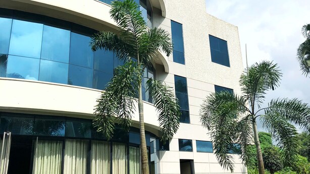 A modern building with large windows and a curved facade, surrounded by potted plants and a well-maintained lawn. - Pai Resorts (Belagavi)