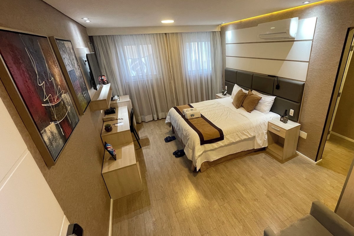A modern hotel room with a large bed, a desk, and a chair. There is a window with curtains, an air conditioning unit, and artwork on the walls.