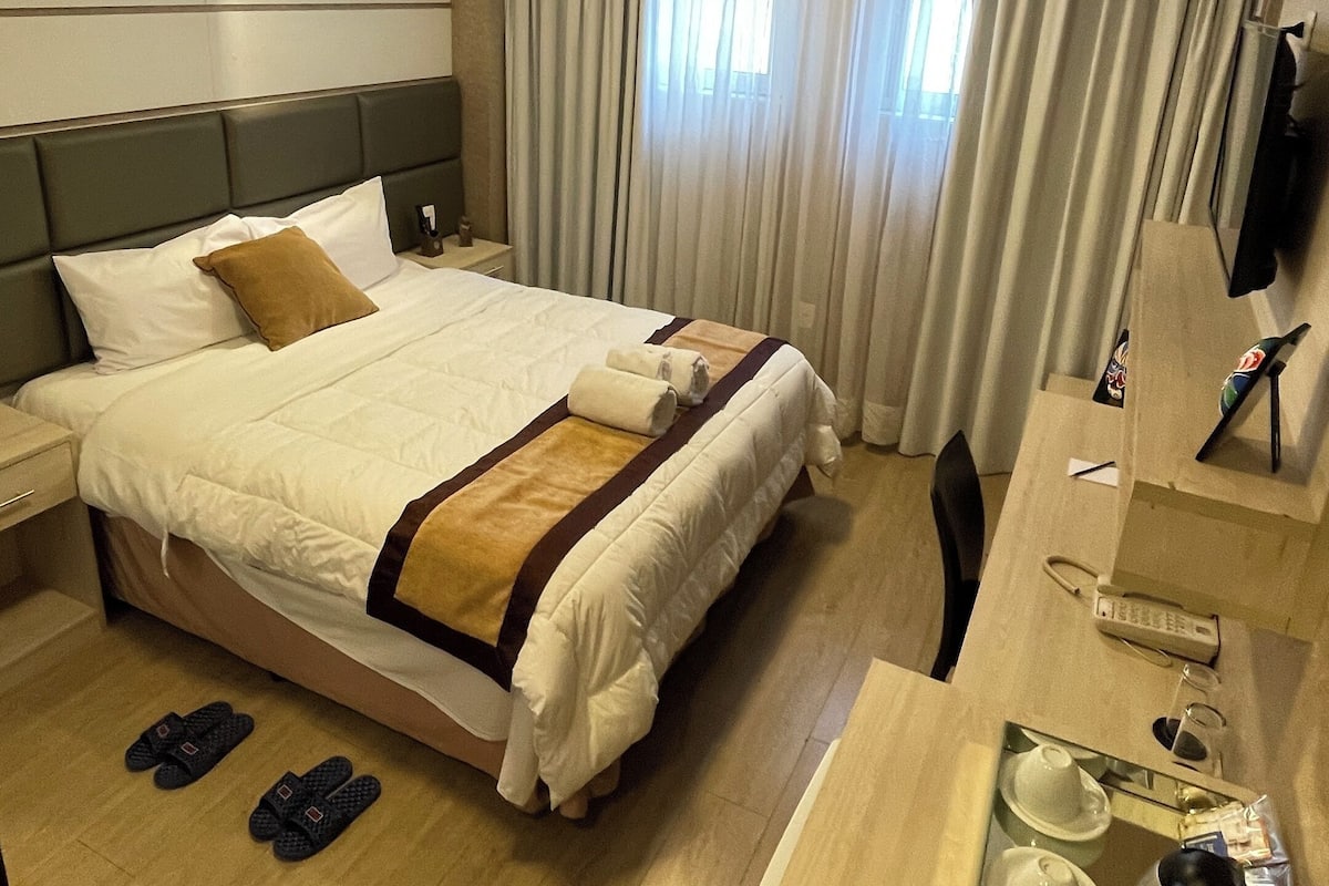 A hotel room with a large bed, a desk with a telephone and a cup, and a window with curtains.