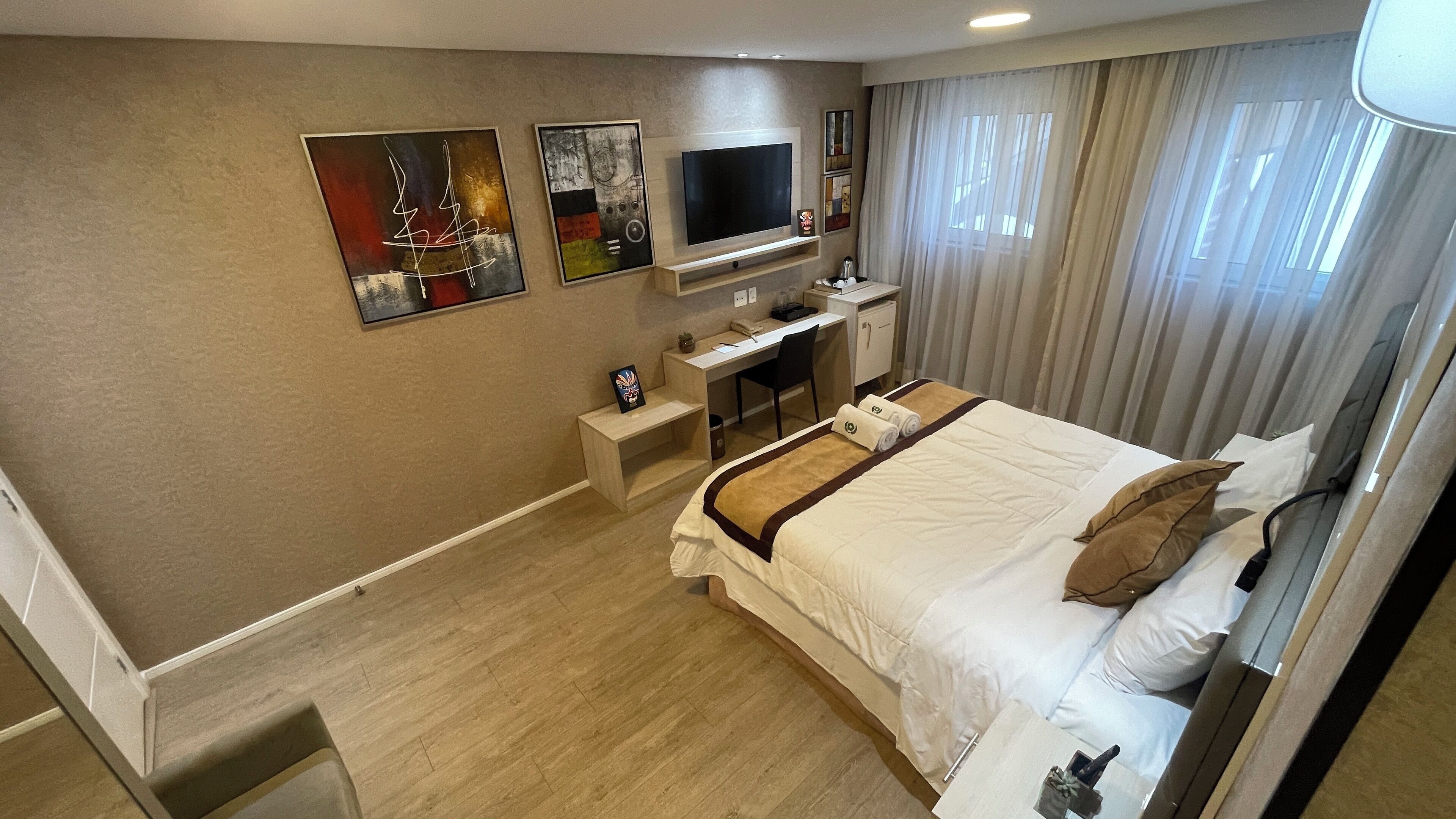 A hotel room with a bed, a desk, a TV, and artwork on the wall.