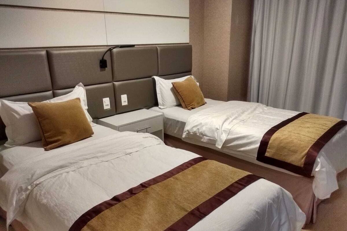 A hotel room with two beds, each with a white bedspread and brown accents, a grey headboard, and a bedside table with a lamp.