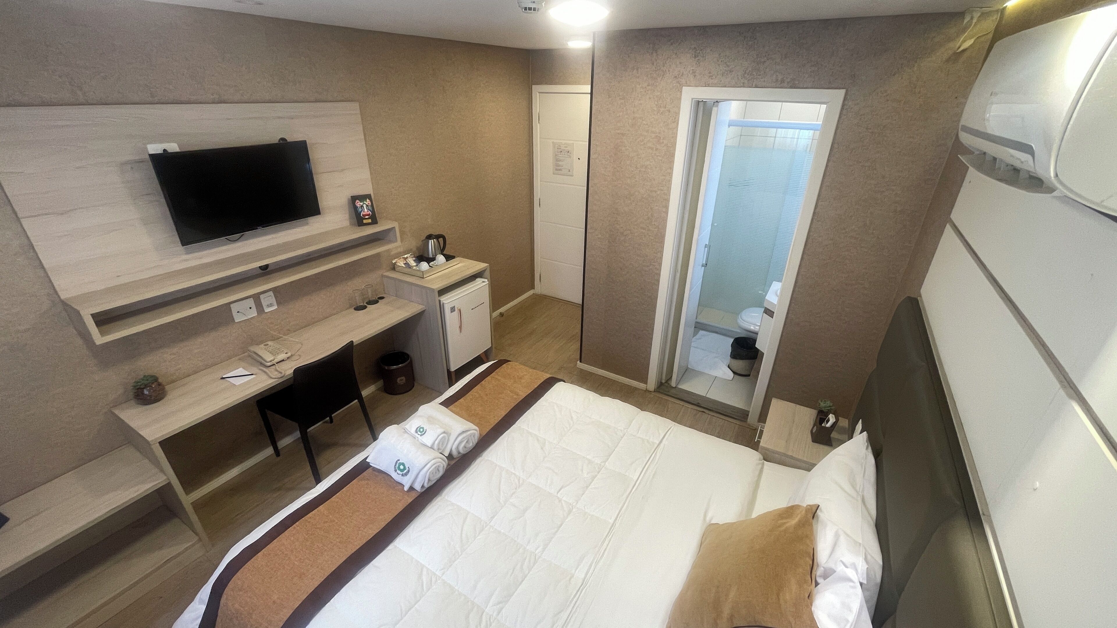 Habitación de hotel con cama, escritorio, televisor, armario y baño visible a través de una puerta abierta.