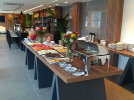 Disposição de buffet com diversos alimentos e um canto de bebidas, rodeado por cadeiras e mesas.