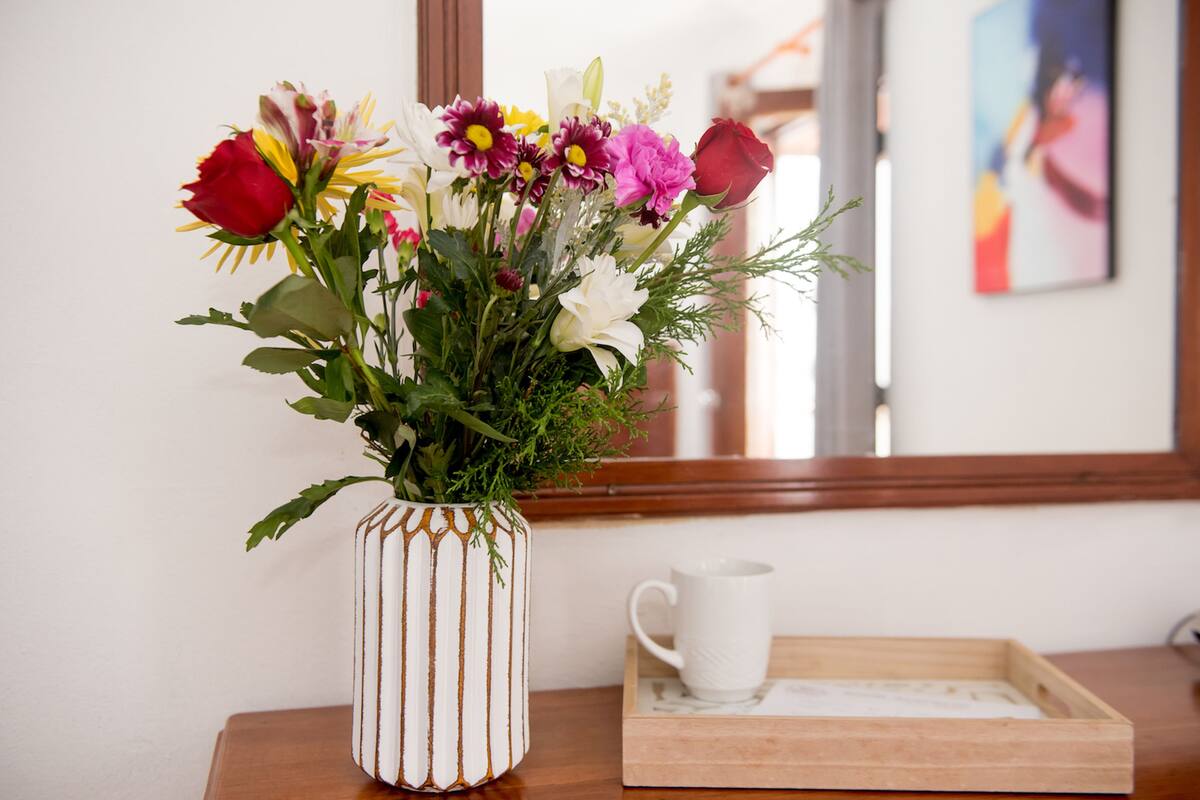 A white vase with vertical lines containing a bouquet of flowers, a white mug, and a wooden tray with a marble pattern on a wooden table.