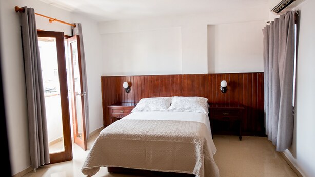 Une chambre d’hôtel avec un lit, une tête de lit en bois, deux tables de chevet et une fenêtre avec des rideaux.