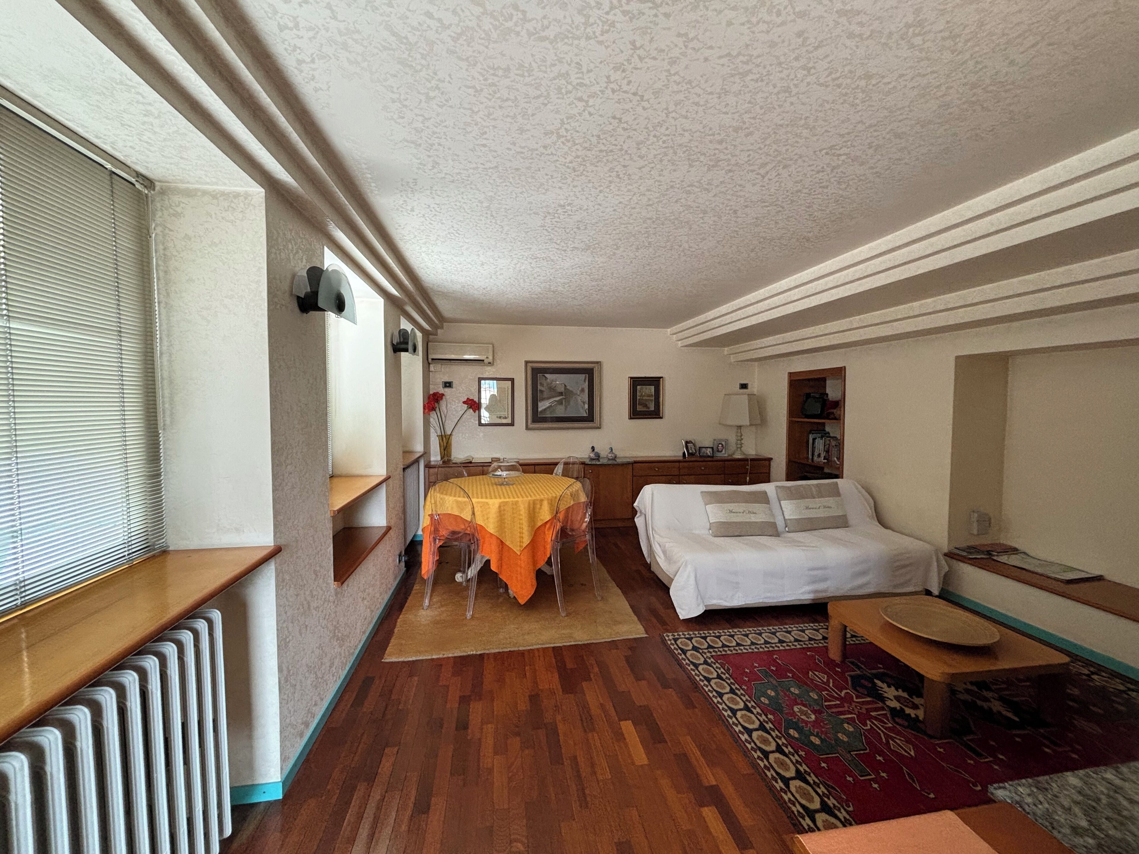 A cozy room with a bed, a small table, a chair, a radiator, a window with blinds, and a wall-mounted air conditioner.