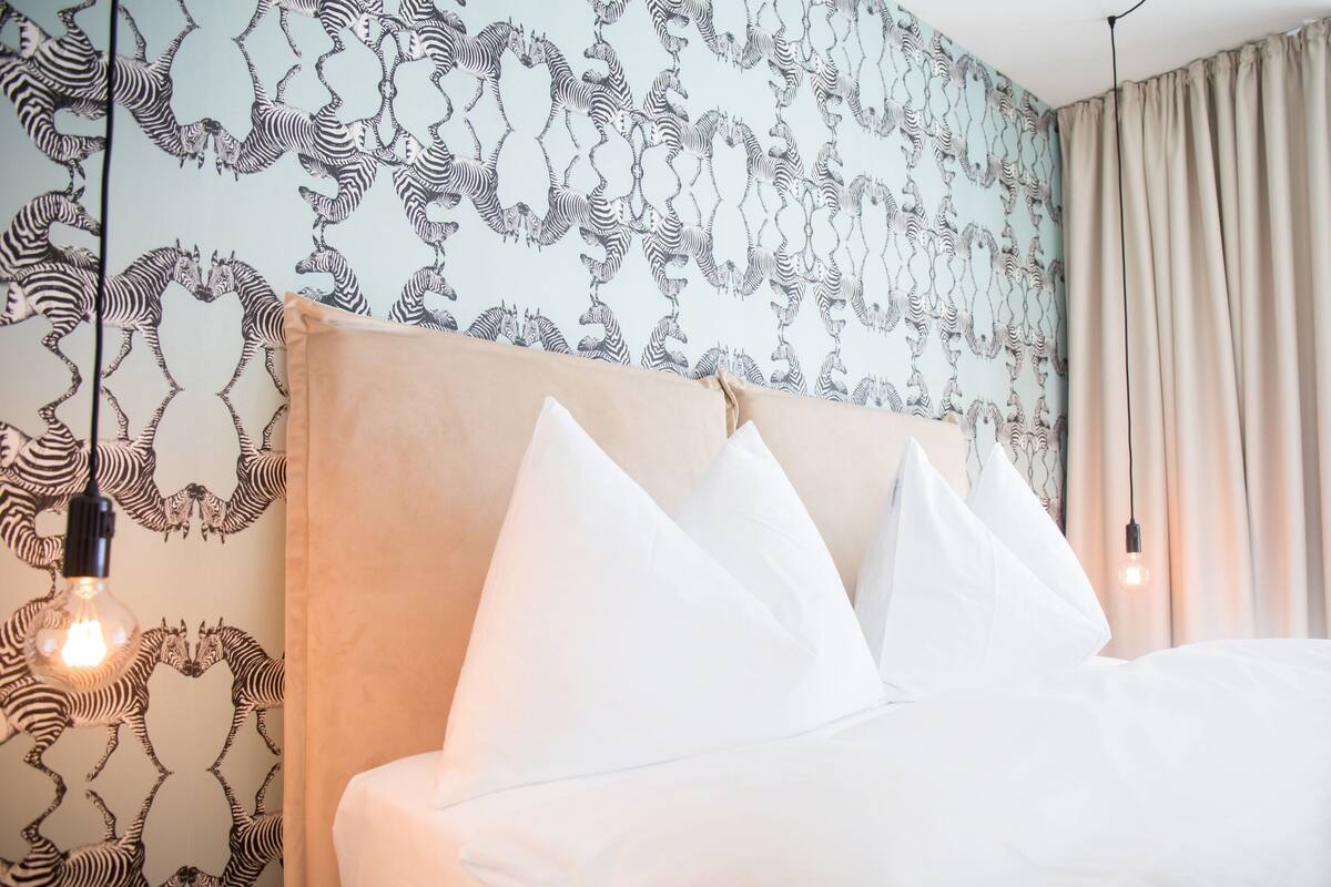 Una cama con almohadas blancas y un cabecero, una pared con papel tapiz estampado con cebra, y una bombilla colgante.
